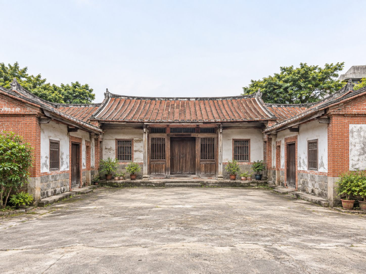 台灣傳統三合院祖厝的正面,中央正廳與兩側護龍圍出前埕,紅磚牆與白灰壁交錯、紅瓦燕尾屋脊,門前擺著幾盆盆栽